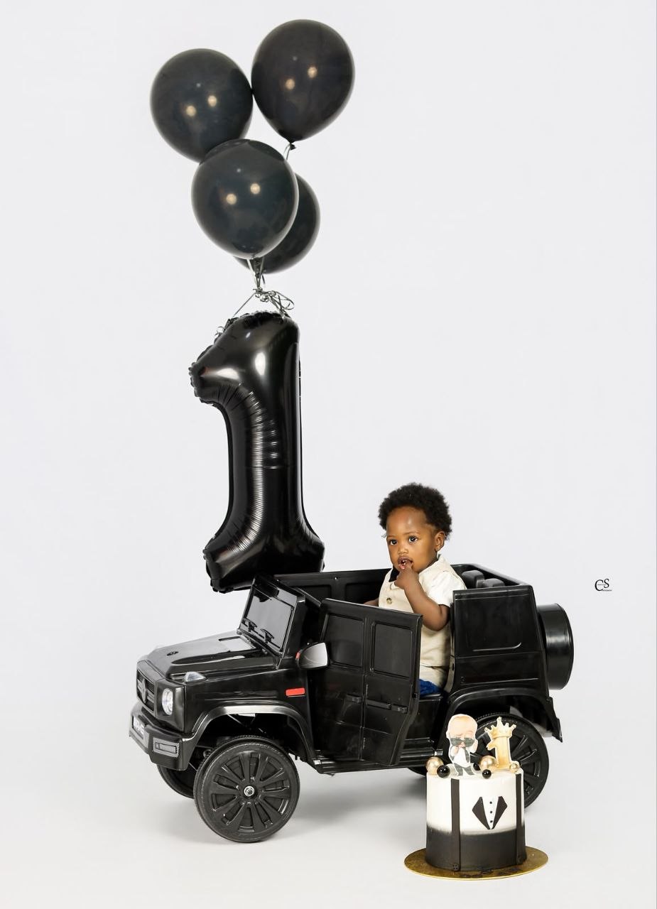Baby boy in a black toy car with birthday balloons